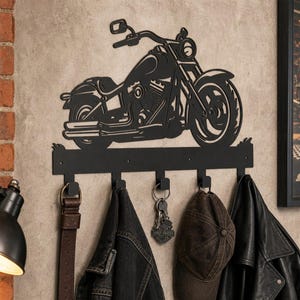 Könnte beinhalten: Eine schwarze Wandgarderobe aus Metall mit der Silhouette eines Motorrads über fünf Haken. An der Garderobe hängen ein brauner Ledergürtel, eine dunkle Jeansjacke, ein Schlüsselbund mit Metallanhänger, eine braune Baseballkappe und eine schwarze Jacke.