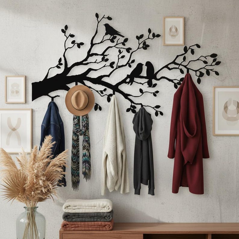 Puede incluir: Un perchero de pared de metal negro con forma de rama de &aacute;rbol con dos p&aacute;jaros. Varios abrigos, un sombrero y una bufanda cuelgan de los ganchos. La pared es de color gris claro y hay grabados art&iacute;sticos enmarcados.