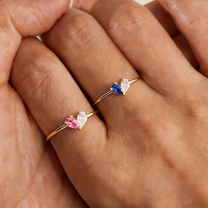 May include: Two gold rings, each featuring a pair of teardrop-shaped gemstones. One ring has a pink and white stone combination, while the other showcases blue and white stones. The rings are displayed on a hand.