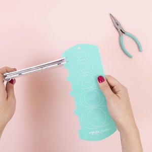 以下が含まれることがあります： 赤いマニキュアを塗った人が持っている、さまざまなサイズの円があるターコイズ色のクラフトテンプレート。小さなシルバーのノートと、ターコイズ色のハンドルのペンチの隣にあります。テンプレートにはcm単位の測定値があります。