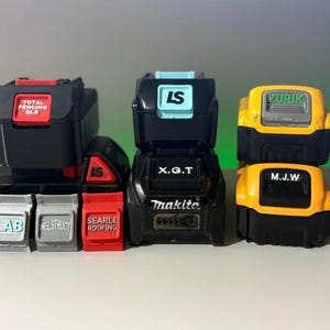 May include: A collection of black and colored power tool battery replicas. The batteries have text labels such as "TOTAL FENCING QLD", "LS", "Makita", and "ZURIK". The colors include black, red, yellow, and light blue.