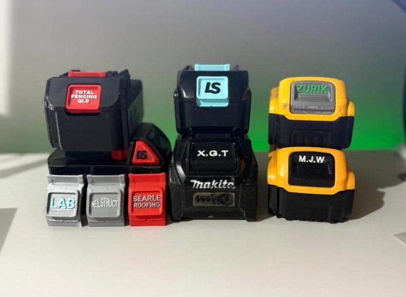 May include: A collection of black and colored power tool battery replicas. The batteries have text labels such as "TOTAL FENCING QLD", "LS", "Makita", and "ZURIK". The colors include black, red, yellow, and light blue.