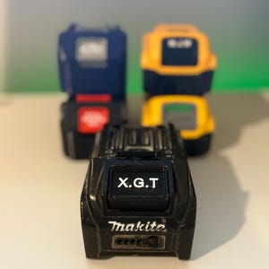 Pode incluir: Bateria preta Makita X.G.T. em primeiro plano, com outras duas baterias no fundo. A bateria X.G.T. tem o texto "X.G.T" e "Makita" impresso. As outras baterias são azuis e amarelas.