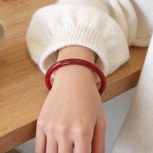 May include: A polished, deep red bangle bracelet. The bracelet, worn on a wrist, contrasts with a white sweater. The circular bracelet's colour is a rich, translucent red, and appears to be made of a polished material.