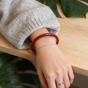 May include: A red bangle bracelet and a silver chain bracelet are worn on a wrist. A silver ring is on the ring finger. The person is wearing a grey jumper. The background includes a wooden surface and a green leaf.