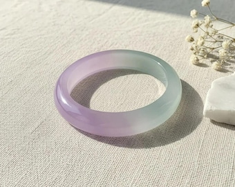 May include: A translucent jade bangle bracelet with a gradient of lavender to pale green. The smooth, polished surface reflects light, showcasing the natural variations in color. The bracelet is displayed on a textured, off-white surface.