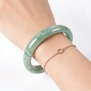May include: A jade bangle bracelet and a gold chain bracelet are displayed on a wrist. The jade bracelet is a light green colour with a smooth, polished surface. The gold bracelet has a delicate chain with a small circular charm.