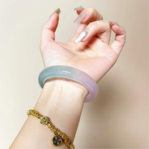 Puede incluir: Un brazalete de jade translúcido en tonos azules y rosas. También se ve una pulsera dorada con detalles turquesa y rojos. Las joyas se muestran en una muñeca.