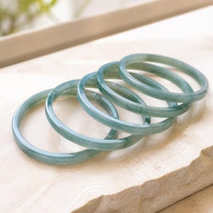 Pode incluir: Uma coleção de cinco pulseiras circulares e lisas, feitas de jade verde-pálido translúcido. Cada pulseira apresenta uma superfície polida e arredondada com variações subtis na intensidade da cor, criando um conjunto coeso de peças de joalharia.
