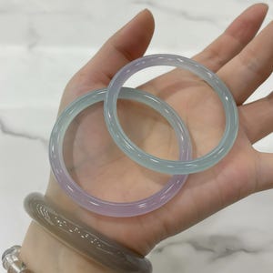 Könnte beinhalten: Zwei durchscheinende Jade-Armreifen in Lavendel- und Meerschaumgrün werden in einer Hand gehalten. Die Armreifen sind glatt und kreisförmig, mit poliertem Finish. Ein dritter, dunklerer Armreif ist am Handgelenk zu sehen.