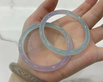 Peut inclure: Deux bracelets en jade translucide, dans des tons de lavande et de vert d'eau, sont tenus dans une main. Les bracelets sont lisses et circulaires, avec une finition polie. Un troisième bracelet, plus foncé, est visible sur le poignet.