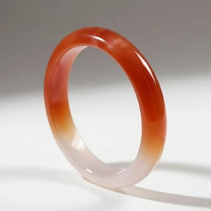 Peut inclure: Un bracelet jonc translucide orange et blanc. Le bracelet a une surface lisse et polie avec un effet de couleur dégradé, passant d'un orange vif à un blanc doux. Le bijou est présenté sur une surface blanche.
