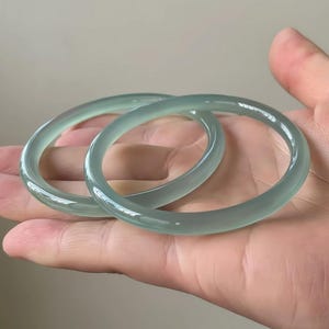 Puede incluir: Dos brazaletes de jadeíta translúcidos de color verde pálido. Las pulseras lisas y pulidas son redondas y parecen estar hechas de una sola pieza de jade. Los brazaletes se muestran sobre un fondo neutro.