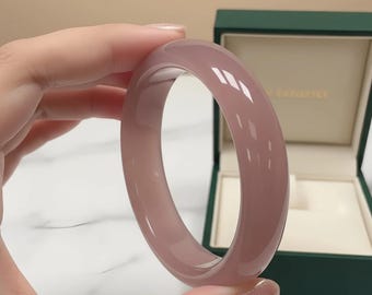 May include: A translucent, mauve-colored bangle bracelet. The smooth, polished surface reflects light, creating highlights. The bracelet is displayed near a green jewelry box with a gold-colored interior.