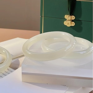 Può includere: Tre braccialetti di giada traslucida bianco-avorio sono esposti su una superficie bianca. I braccialetti lisci e arrotondati sono impilati, mostrando la loro finitura lucida. Sullo sfondo, una scatola verde scuro con dettagli dorati e un nastro di velluto.