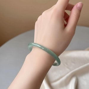 May include: A pale green jade bangle bracelet. The smooth, rounded bracelet is a light green colour and is worn on a wrist. The bracelet is a simple, elegant piece of jewellery.