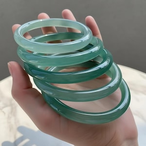 May include: A collection of six translucent jade bangles in varying shades of green. The smooth, polished bracelets are stacked in a hand, showcasing their circular design and glossy finish. The jade appears to have a natural, organic quality.