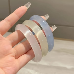 Pode incluir: Quatro pulseiras translúcidas em cores pastel: rosa claro, creme e azul claro. As pulseiras são lisas e arredondadas, com um acabamento brilhante. As pulseiras estão empilhadas, mostrando seus diferentes tons.