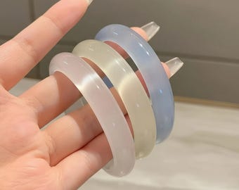 May include: Four translucent bangles in pastel colors: light pink, cream, and light blue. The bracelets are smooth and rounded, with a glossy finish. The bangles are stacked together, showcasing their different hues.