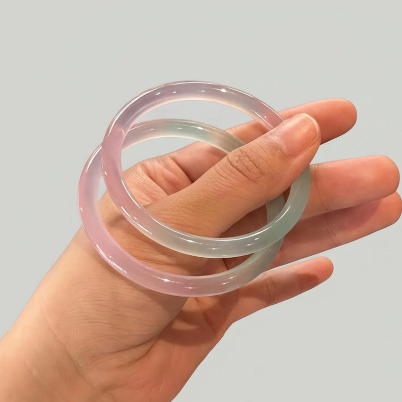 May include: Two translucent jade bangles in shades of pink and green are held in a hand. The smooth, polished bracelets are circular and appear to be made of a natural stone. The background is a solid, neutral color.