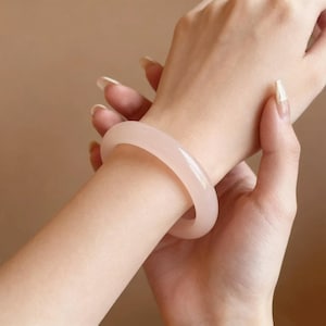 Könnte beinhalten: Ein hellrosa Jade-Armreif. Der glatte, polierte Armreif wird am Handgelenk getragen und zeigt seine sanfte Farbe und runde Form. Der Hintergrund ist ein neutrales Beige.