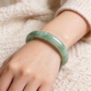 May include: A light green jade bangle bracelet. The smooth, polished bracelet is worn on a wrist, with a cream-coloured sweater in the background. The jade has a marbled appearance with varying shades of green.