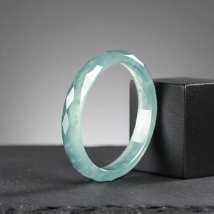 Peut inclure: Un bracelet jonc en jade bleu clair avec un design à facettes. Le bracelet est d'une couleur vert pâle translucide et est présenté sur une surface sombre. Le bracelet est de forme circulaire.