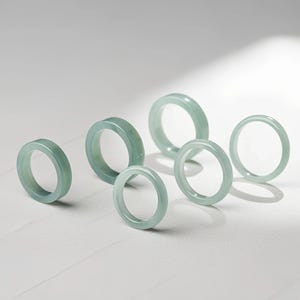 May include: Six jade rings of varying sizes are displayed against a white background. The rings have a pale green hue and a smooth, polished surface. The rings are circular and appear to be made of a translucent stone.