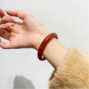 May include: A polished, deep red bangle bracelet. The smooth, circular bracelet is worn on a wrist, with a neutral background. The bracelet is approximately 2 cm wide. The wrist is covered by a tan, fuzzy sleeve.