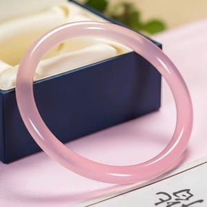 May include: A pale pink bangle bracelet, circular in shape and made of a smooth, polished material. It is displayed on a pink surface next to a dark blue jewellery box.