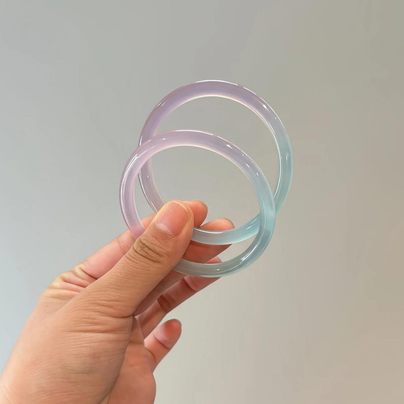 May include: Two translucent, round bangles in shades of lavender and seafoam green. The bracelets are held in a hand, showcasing their smooth, polished surface. The light reflects off the curved surfaces, creating a soft, ethereal glow. These accessories are perfect for adding a touch of color.