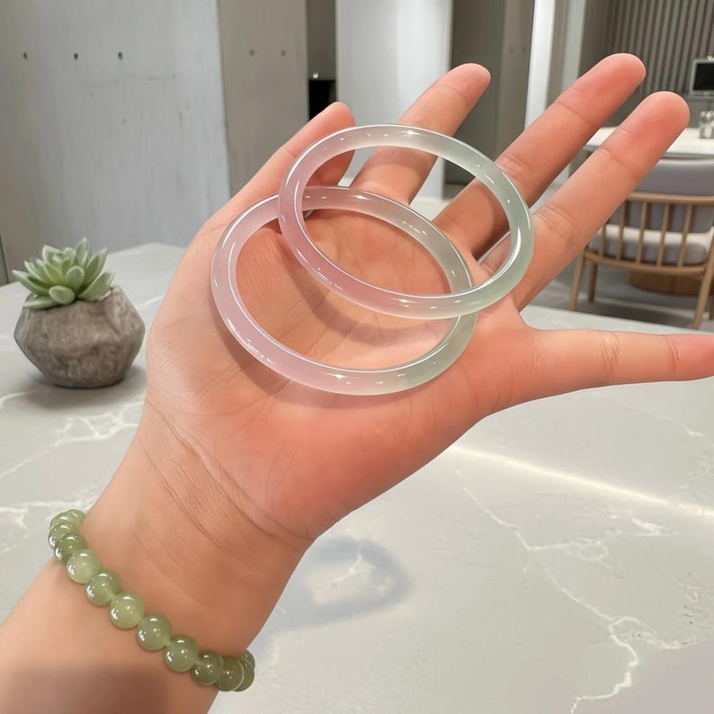 May include: Two translucent jade bangles, one with a gradient of pink and green hues, are held in a hand. A jade bead bracelet is worn on the wrist. The background includes a succulent plant in a stone pot.