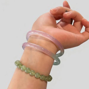 May include: A wrist adorned with two translucent, pastel-colored bangles in shades of pink and blue, and a beaded bracelet of green gemstones. The bracelets are worn on a light-skinned arm against a plain background.