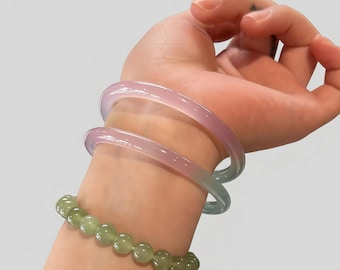 May include: A wrist adorned with two translucent, pastel-colored bangles in shades of pink and blue, and a beaded bracelet of green gemstones. The bracelets are worn on a light-skinned arm against a plain background.