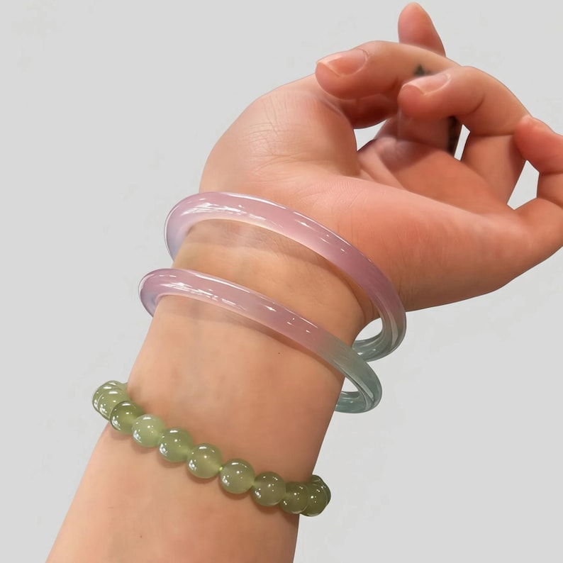 May include: A wrist adorned with two translucent, pastel-colored bangles in shades of pink and blue, and a beaded bracelet of green gemstones. The bracelets are worn on a light-skinned arm against a plain background.