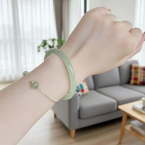 May include: A pale green jade bangle and a gold-coloured open cuff bracelet with a jade bead. The bracelets are shown on an arm, highlighting the jewellery's colour and design. The background includes a sofa and a wooden table.