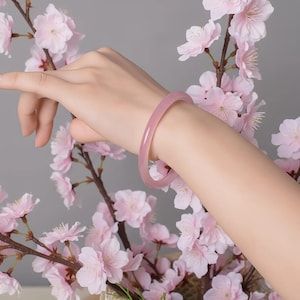 May include: A pale pink, translucent bangle bracelet. The bracelet is worn on a wrist, with a backdrop of pink cherry blossoms. The bracelet appears to be made of a smooth, polished material.