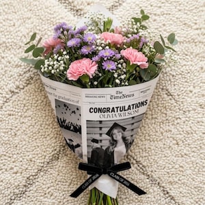 May include: A bouquet of pink carnations, purple asters, and white baby's breath wrapped in newspaper. The newspaper reads "Congratulations Olivia Wilson" with a photo of a graduate. A black ribbon with "Class of 2016" is tied around the stems.