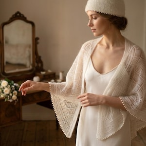 May include: A cream-colored, open-weave shawl with a delicate pattern and beaded trim. The shawl is draped over a white dress and paired with a matching hat. The image is set in a room with a vintage vanity and a bouquet of white flowers.
