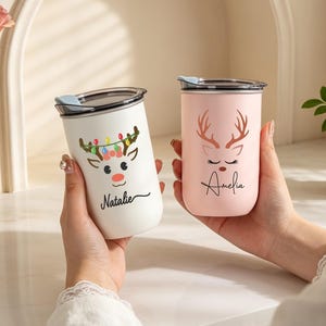 May include: Two cylindrical tumblers with lids, one white and one light pink. The white tumbler features a reindeer graphic with colourful lights and the name Natalie. The pink tumbler displays a reindeer graphic with antlers and the name Amelia.