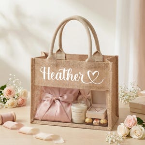 Pode incluir: Uma sacola de juta com o nome "Heather" e um coração. A sacola tem um painel frontal transparente revelando um roupão rosa, uma vela, joias e bombons. A sacola está em uma superfície clara com flores.