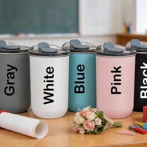 May include: Five insulated tumblers in gray, white, blue, pink, and black, each with a matching lid and the color name printed vertically. A rolled diploma, flowers, and a gift box are on the wooden surface.