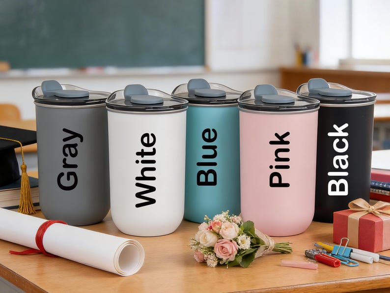 May include: Five insulated tumblers in gray, white, blue, pink, and black, each with a matching lid and the color name printed vertically. A rolled diploma, flowers, and a gift box are on the wooden surface.
