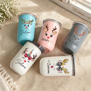 Könnte beinhalten: Fünf Edelstahlbecher mit Deckel, verziert mit Rentiergrafiken und personalisierten Namen. Die Becher sind hellblau, rosa, grau und weiß und zeigen verschiedene Geweihdesigns, rote Nasen und Schleifen. Die Namen sind Salome, Natalie und Amelie.