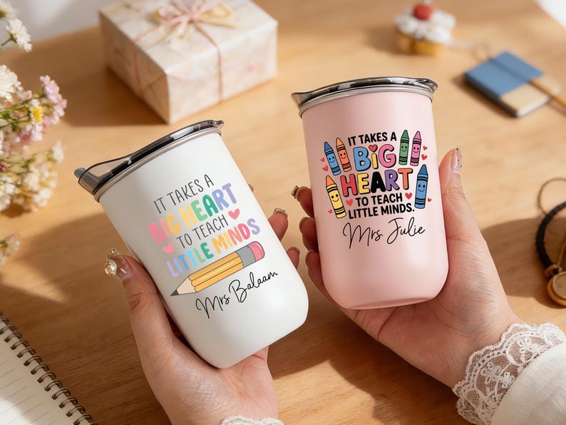 May include: Two stainless steel tumblers, one white and one pink, with black lids. The white tumbler has the text "IT TAKES A BIG HEART TO TEACH LITTLE MINDS" and a pencil graphic. The pink tumbler has the same text and crayon graphics.