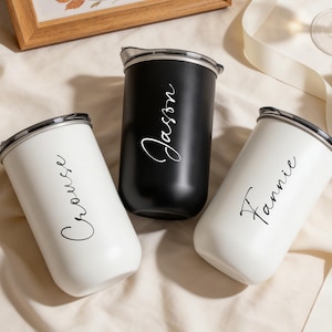 Op de afbeelding: Drie cilindrische bekers met doorzichtige deksels, twee witte en één zwarte. Elke beker is voorzien van een aangepaste naam in cursief schrift: Crouse op een witte beker, Fannie op de andere witte beker en Jason op de zwarte beker.