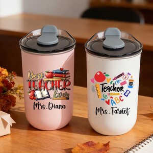 May include: Two insulated tumblers with black lids and gray flip-top closures. One is pink with "Best Teacher Ever" text and the other is white with a heart-shaped teacher graphic. Both have teacher's names.