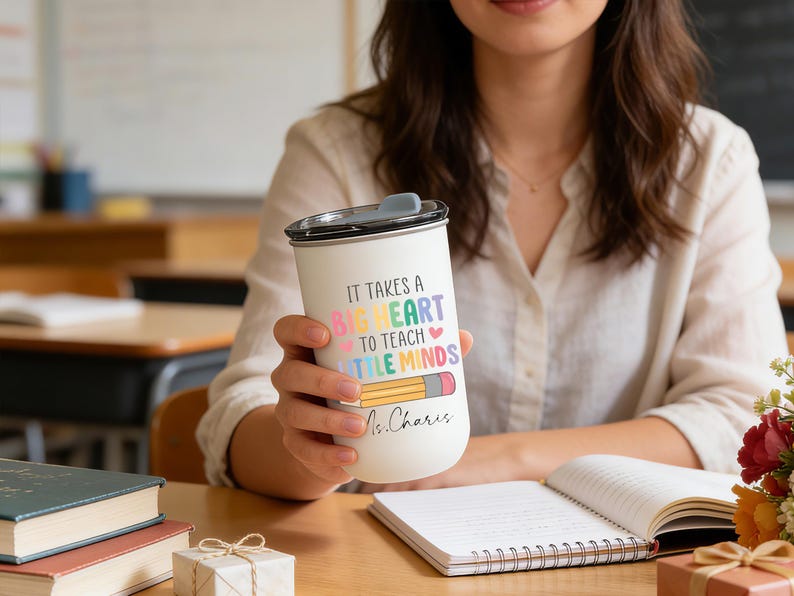 May include: A white travel mug with a black lid, featuring the text "IT TAKES A BIG HEART TO TEACH LITTLE MINDS" in colorful letters and a pencil illustration. The mug is personalized with the name "Ms. Charis".