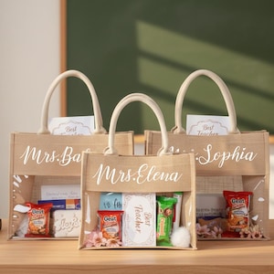 May include: Three beige jute tote bags with clear front panels, each personalized with a teacher's name in white script. The bags are filled with gifts, including snacks and cards, and have cream-colored handles. A mug and books are on the table.