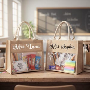 May include: Two beige burlap tote bags with clear front pockets, filled with treats and school supplies. One bag says "Mrs. Lana" and the other says "Mrs. Sophia". The bags are on a wooden desk in a classroom setting.